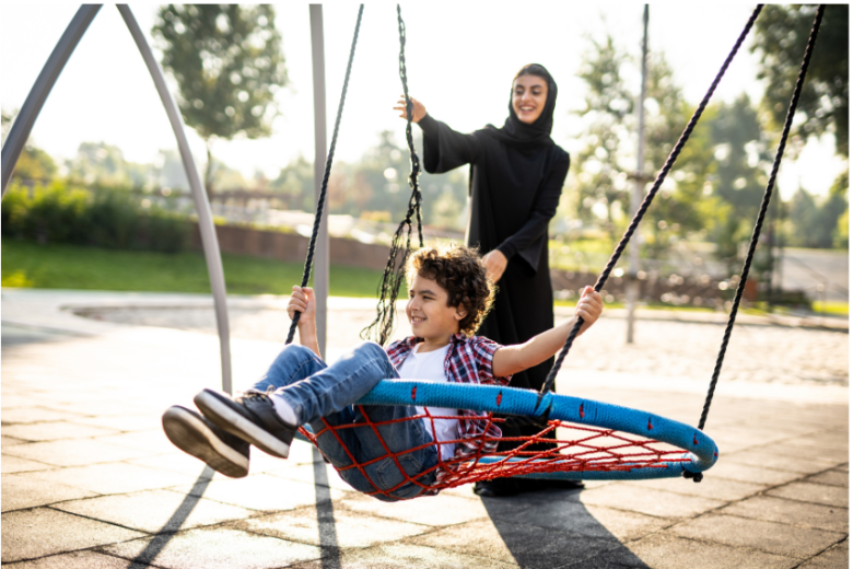 Representing long-term fostering - a woman wearing a hijab and smiling in the sunshine pushes a boy on a nest swing in a park